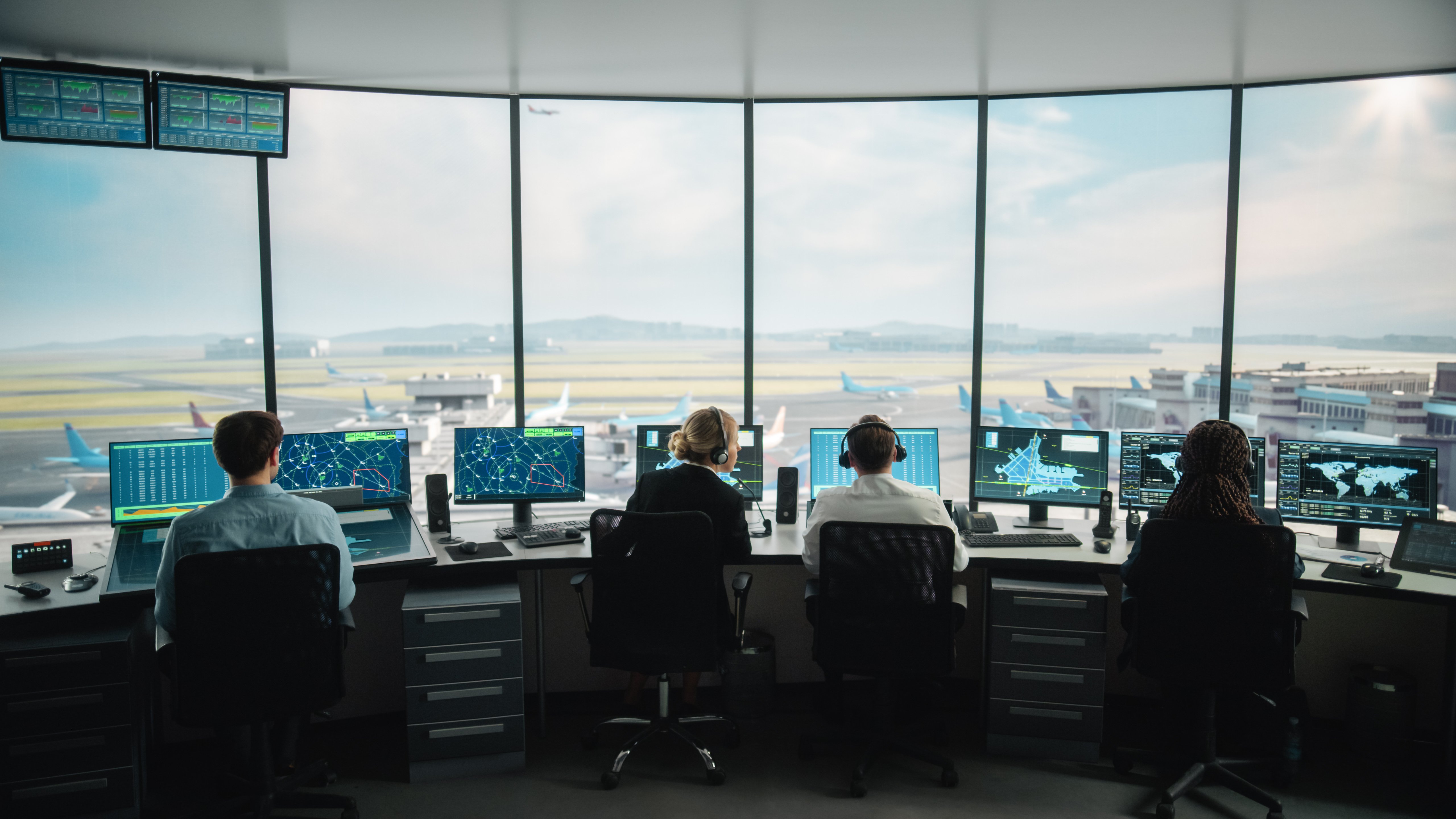 Vier Fluglotsen sitzen an Arbeitsplätzen in einem Flughafenkontrollturm und überwachen jeweils mehrere Bildschirme, auf denen Radar, Flugwege, Flughafenkarten und globale Daten angezeigt werden. Große Panoramafenster bieten einen klaren Blick auf die Start- und Landebahn, die rollenden Flugzeuge und die Terminalgebäude. Das Team koordiniert die Flugbewegungen in einer hochtechnisierten, sicheren Umgebung.