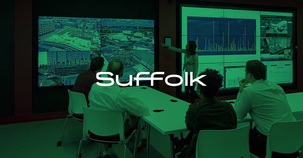 Stehender Mitarbeiter präsentiert auf einer Videowand, 4 Mitarbeiter sehen sich die Präsentation an einem Tisch im Suffolk's L.A. Smart Lab mit grünem Overlay und Logo an
