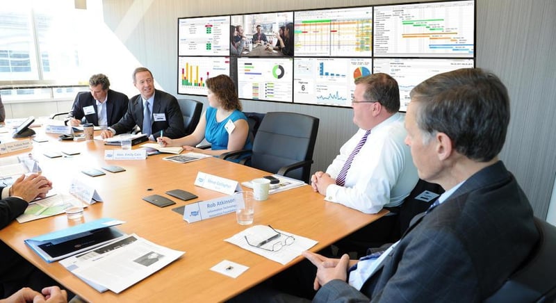 5 Mitarbeiter diskutieren an einem Tisch in einem Besprechungsraum mit einer Videowand, auf der Daten visualisiert werden