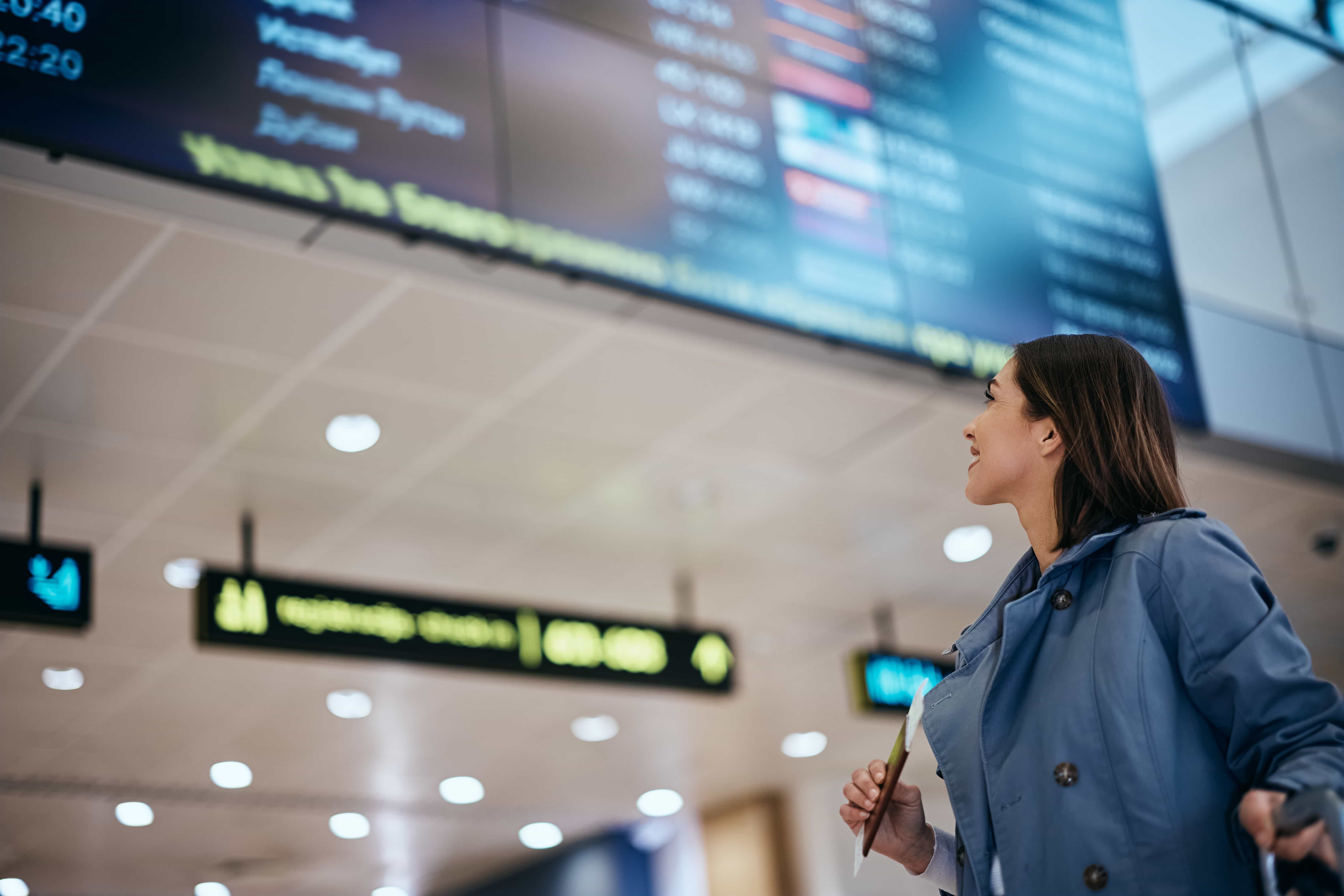  Eine Frau in einer hellblauen Jacke und einem weißen Hemd blickt auf eine große Abflugtafel in einem Flughafen. Sie lächelt leicht und hält in ihrer rechten Hand etwas, das ein Pass oder ein Ticket zu sein scheint. Auf der Abflugtafel, die unscharf ist, werden verschiedene Fluginformationen angezeigt. Im Hintergrund ist auch die beleuchtete Decke des Flughafens zu sehen.