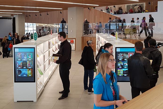 Viele Menschen gehen in einer Bibliothek umher und sehen sich Regale mit digitaler Beschilderung an.