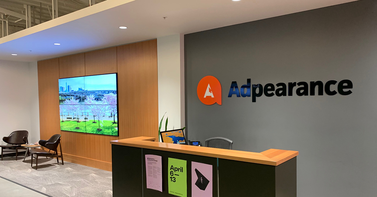 Leeres Foyer von Adpearance, mit einer von Userful betriebenen Videowand, die hinter einer Sitzecke Fotos zeigt, und einem Empfangstresen mit dem Adpearance-Logo an der Wand dahinter