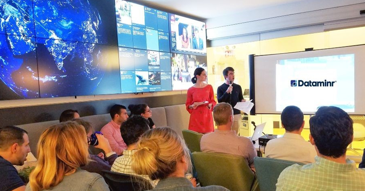 Dataminr-Mitarbeiter in einem Besprechungsraum, gegenüber einem Projektor und neben einer Videowand mit sozialen Medien und aktuellen Nachrichten