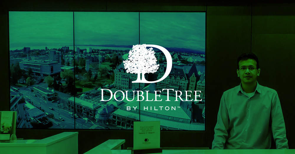 DoubleTree by Hilton-Rezeptionist und 3-Panel-Videowand hinter ihm, die Wahrzeichen von Victoria, Kanada mit grünem Overlay und Logo zeigt