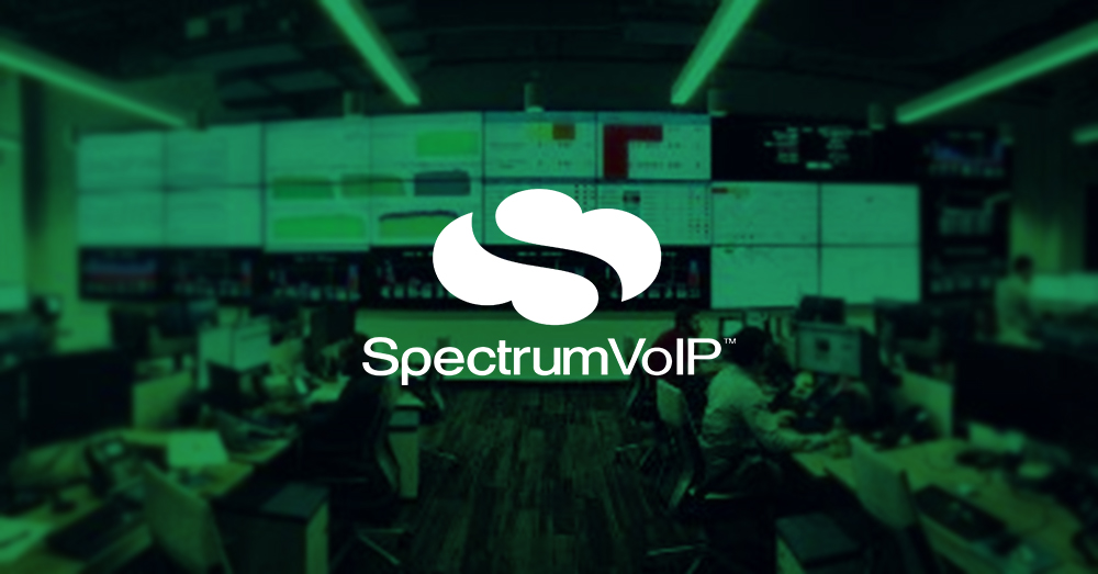 Leeres SpectrumVoIP Network Operations Center mit vielen Arbeitsplätzen und einer größeren Videowand, die Daten-Dashboards mit grünem Overlay und Logo anzeigt