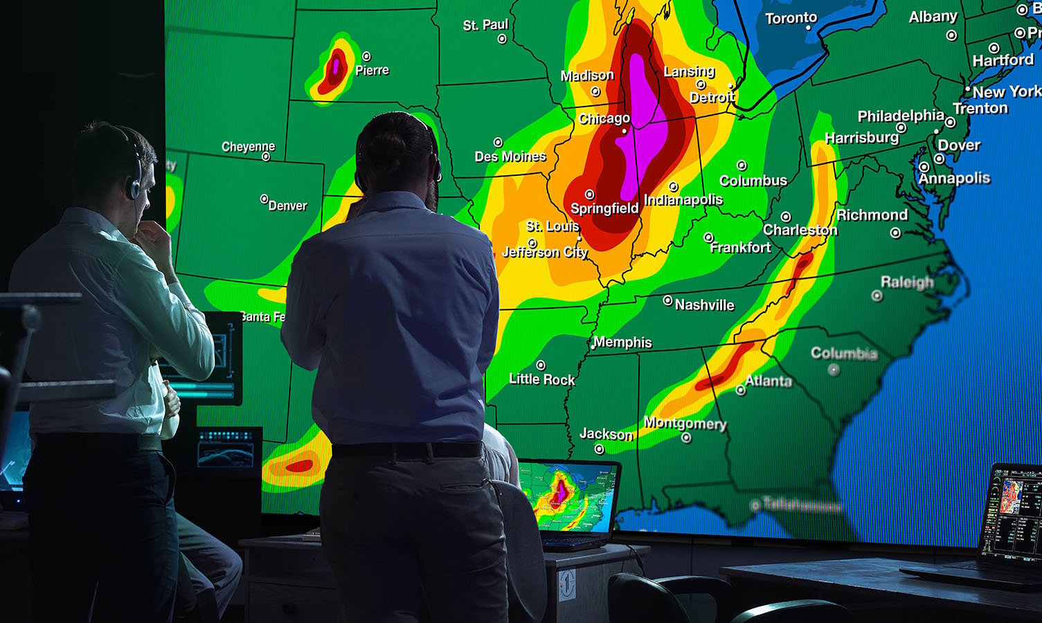2 Männer in einem Kontrollraum für die Wetterüberwachung, die auf eine große Videowand mit einer Wetterkarte der USA schauen